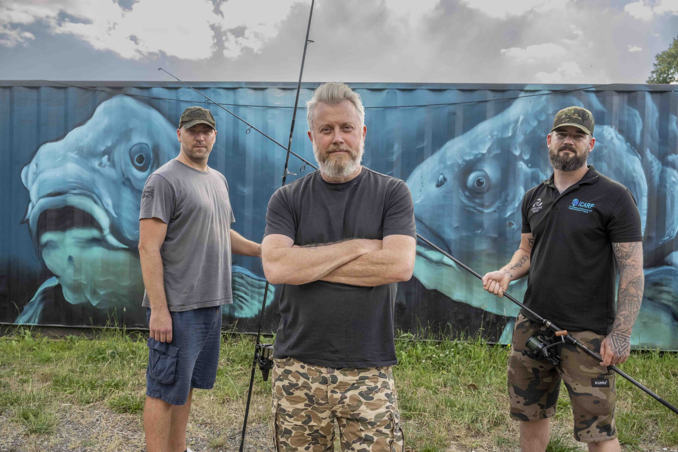 A group of three people holding fishing rods standing in front of graffiti of carp.