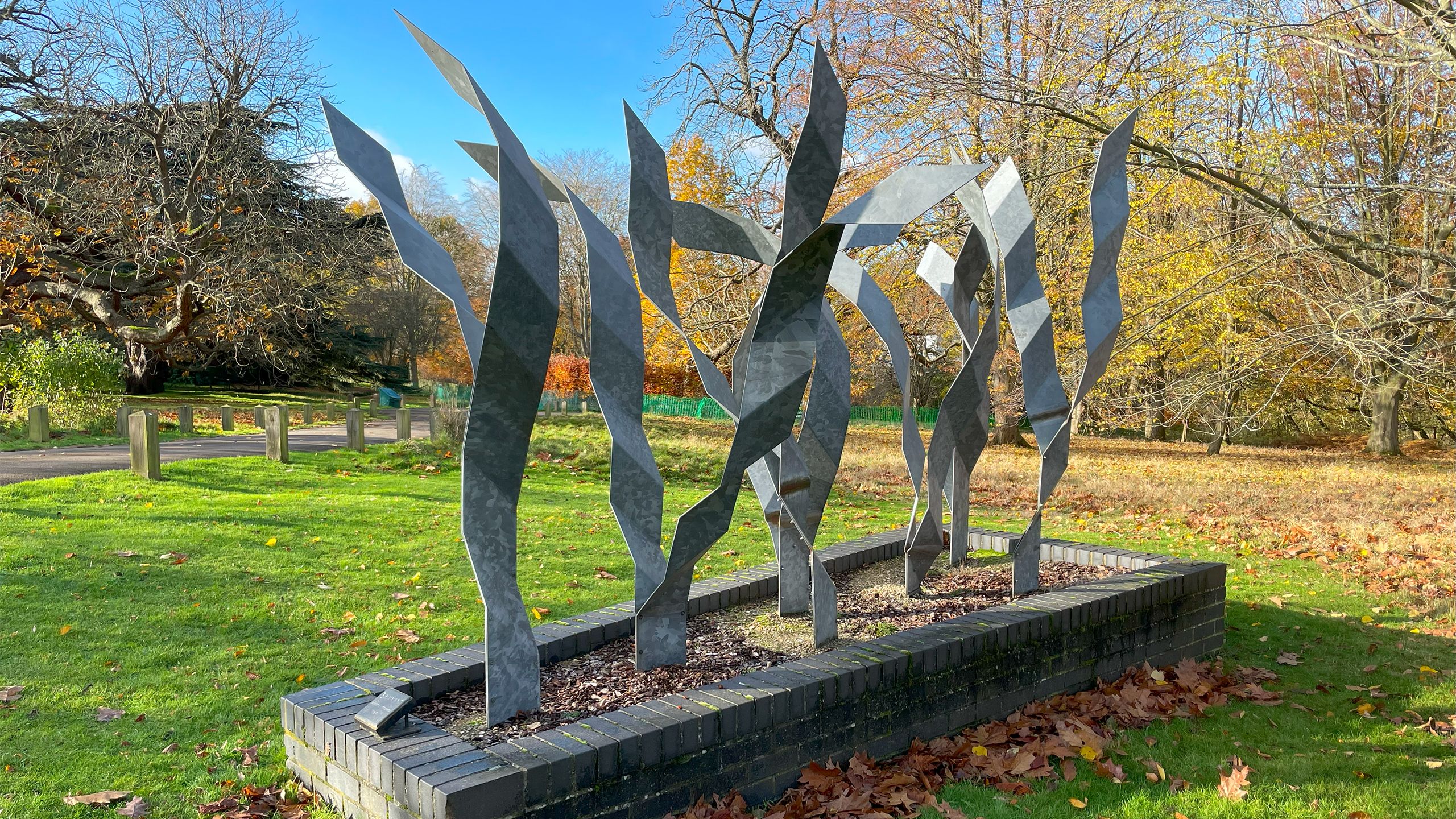 A row of wavy, metallic blades protrude from a brick base among Wivenhoe Park's trees.