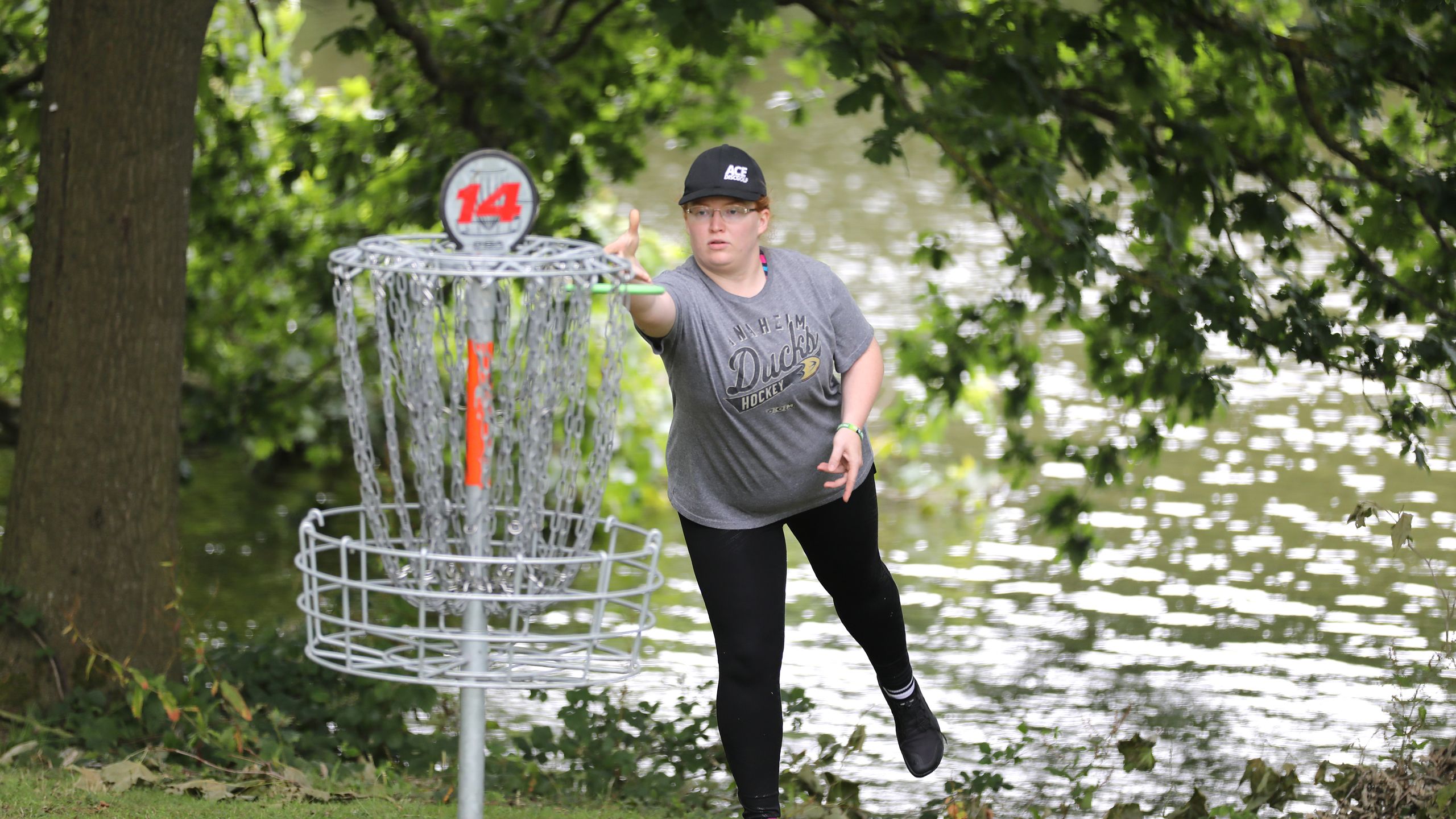 A disc golfer throws a disc towards a net made of chains. Behind them are trees and the lakes in Wivenhoe Park.