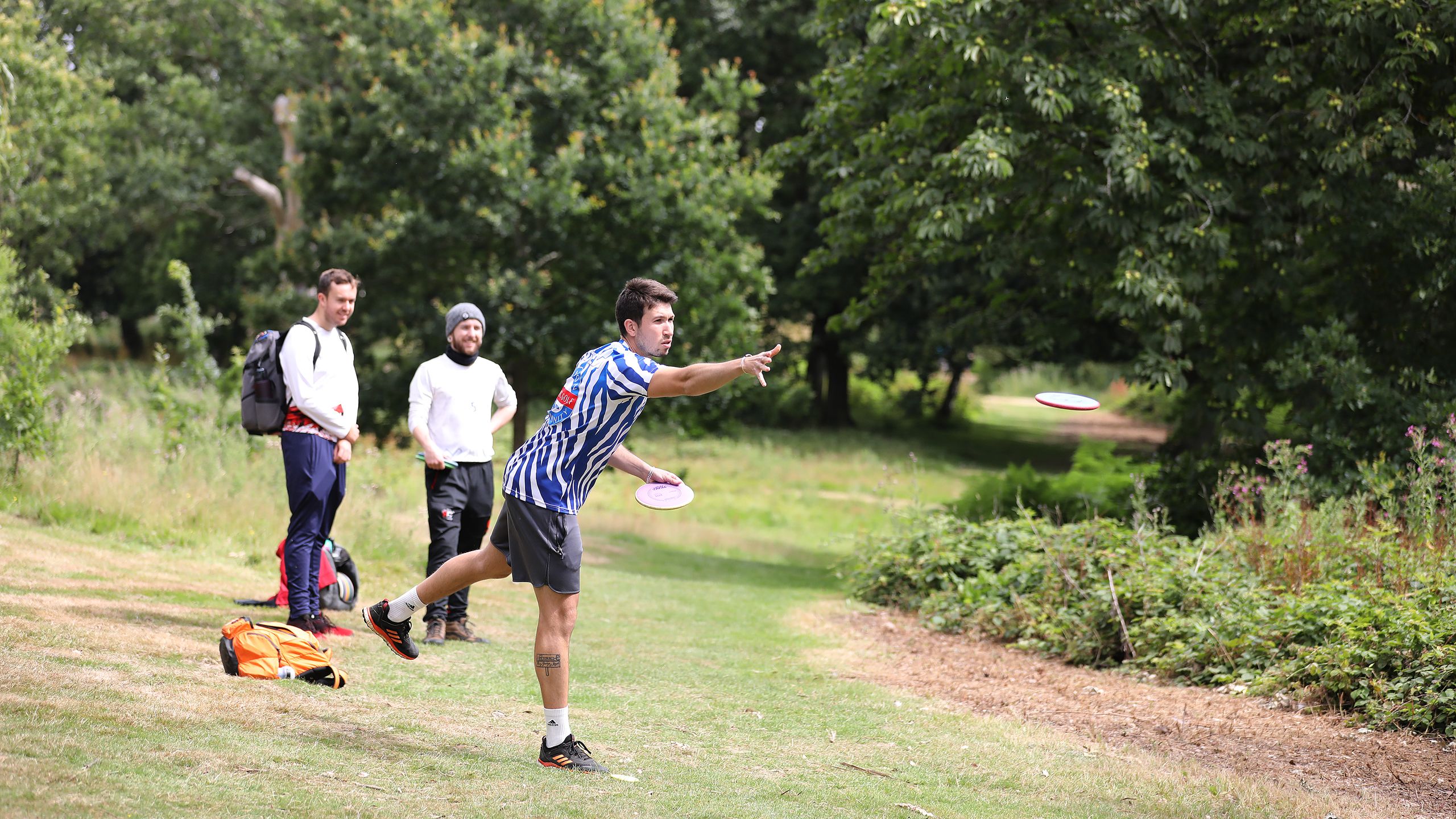 Two spectators watch as a disc golfer throws a disc across the parkland.
