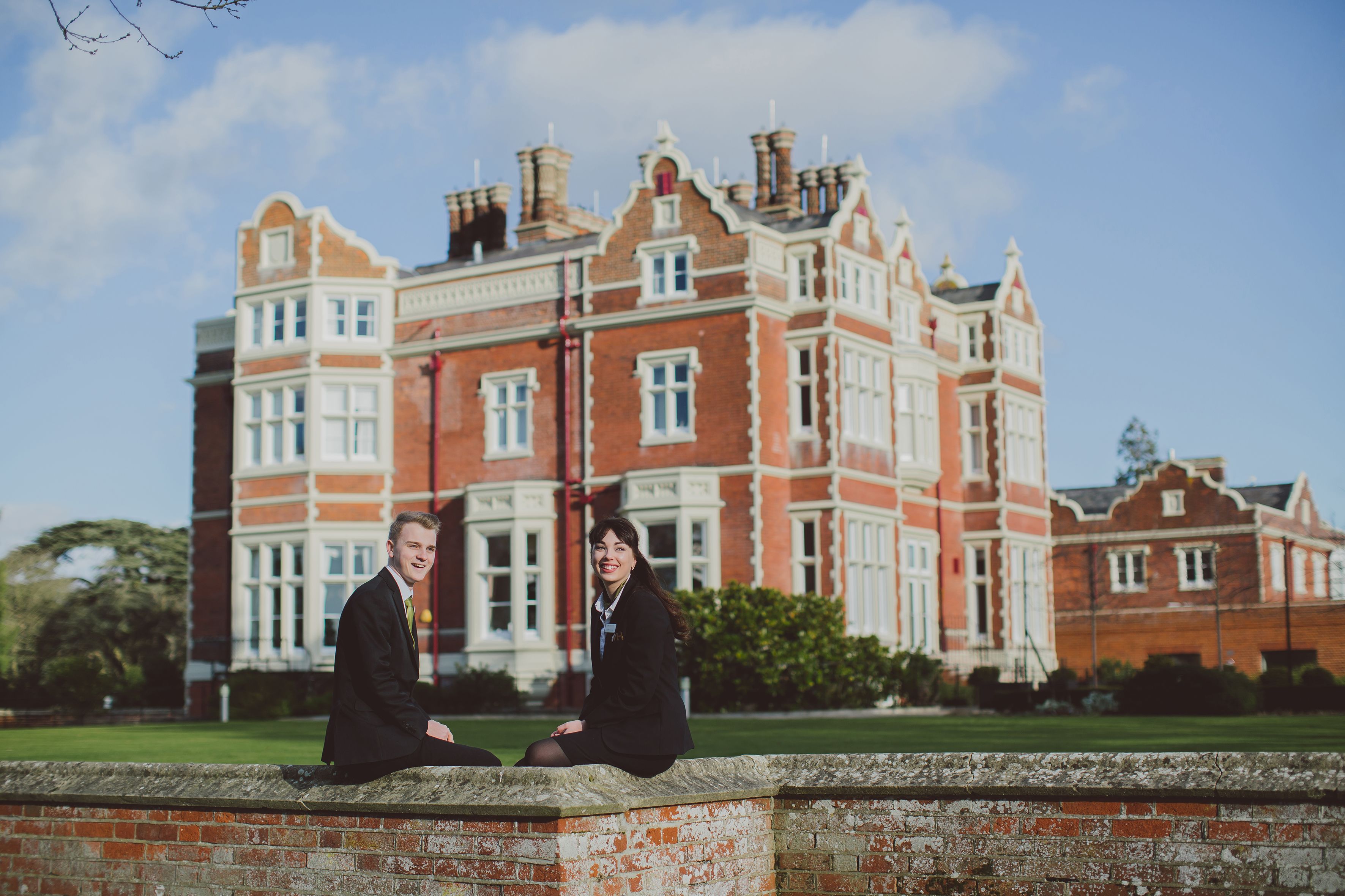 Two young hospitality professionals, smiling, sitting on a low wall outside Wivenhoe House Hotel