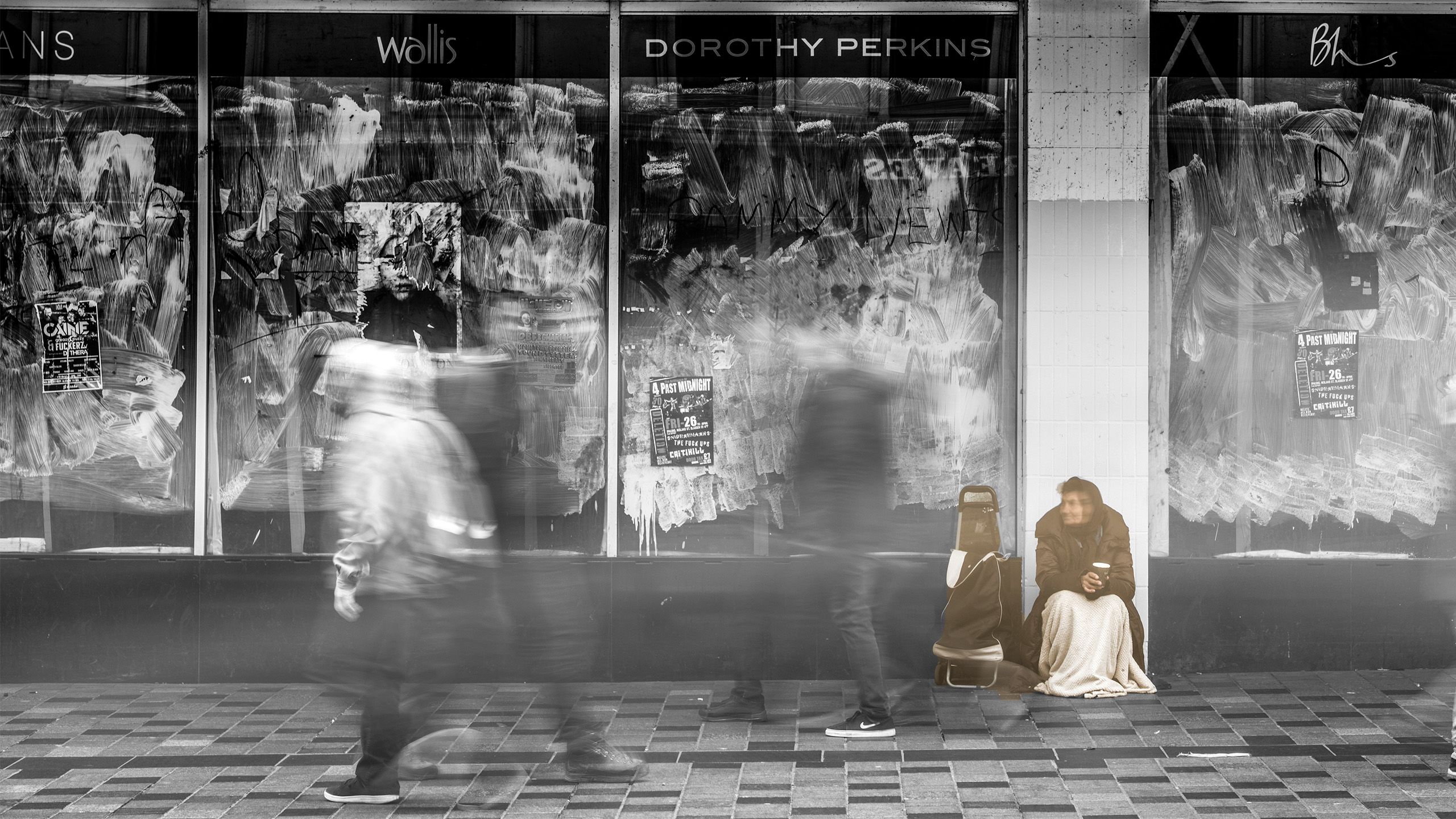 Black and white picture of a homeless lady sat in front of a series of boarded up shops