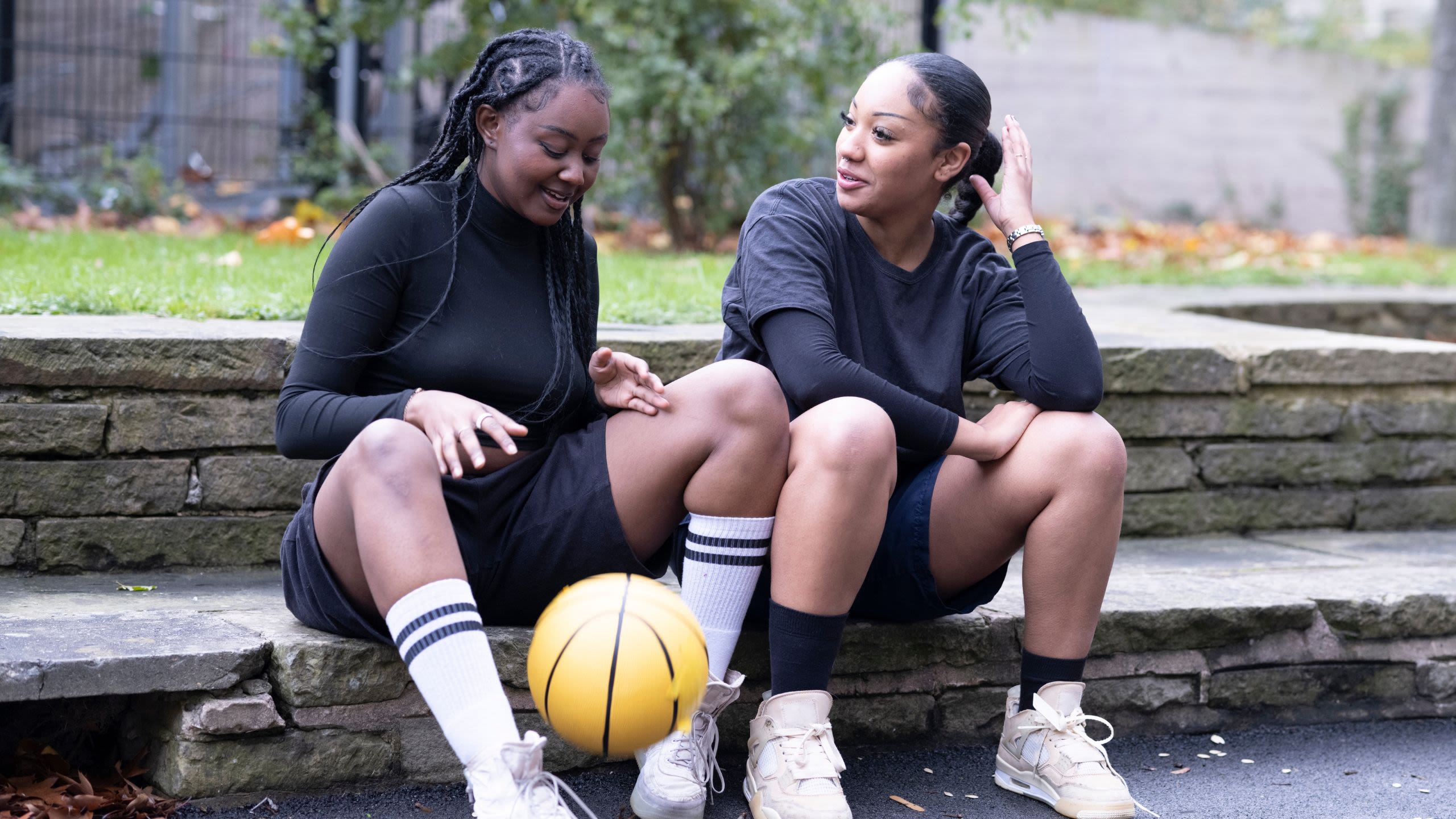 Two teenage girls, wearing black sports clothes, sitting on steps and chatting. One is bouncing a basketball