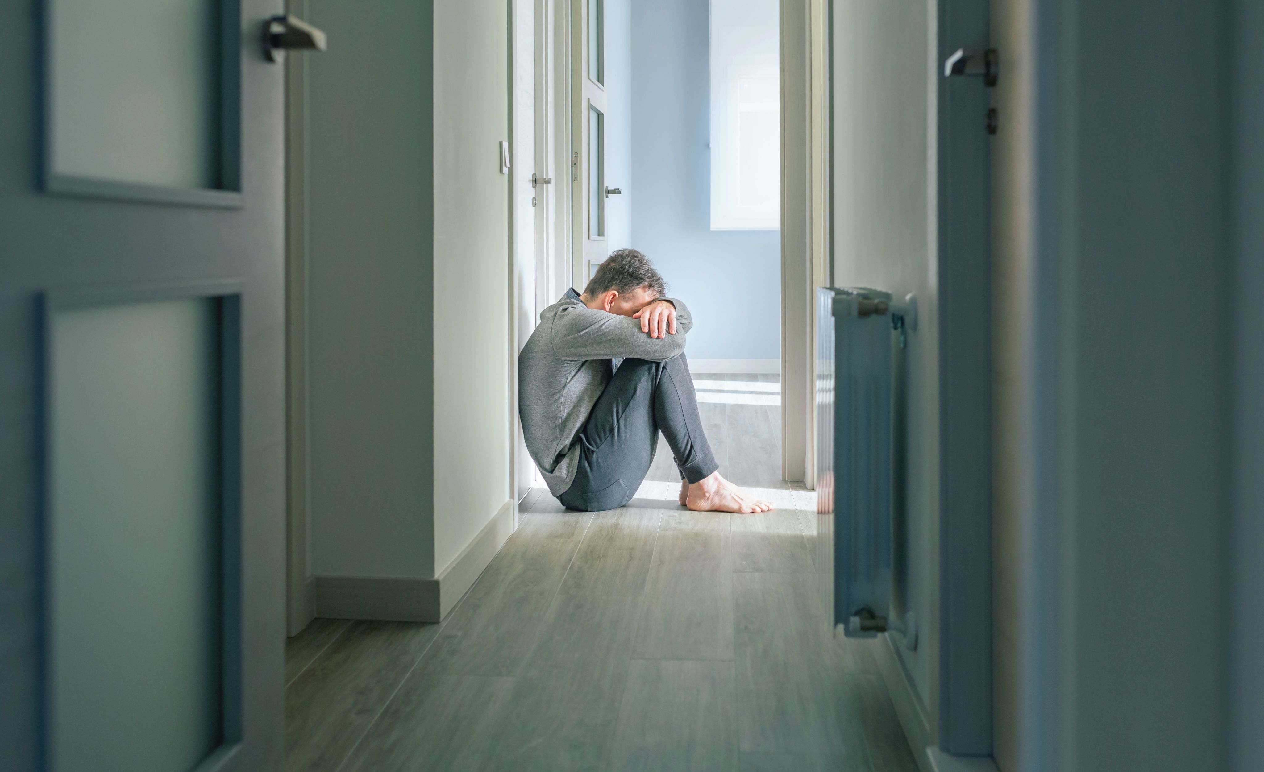A man sits on the floor, at the end of stark looking corridor, holding his head in his hands