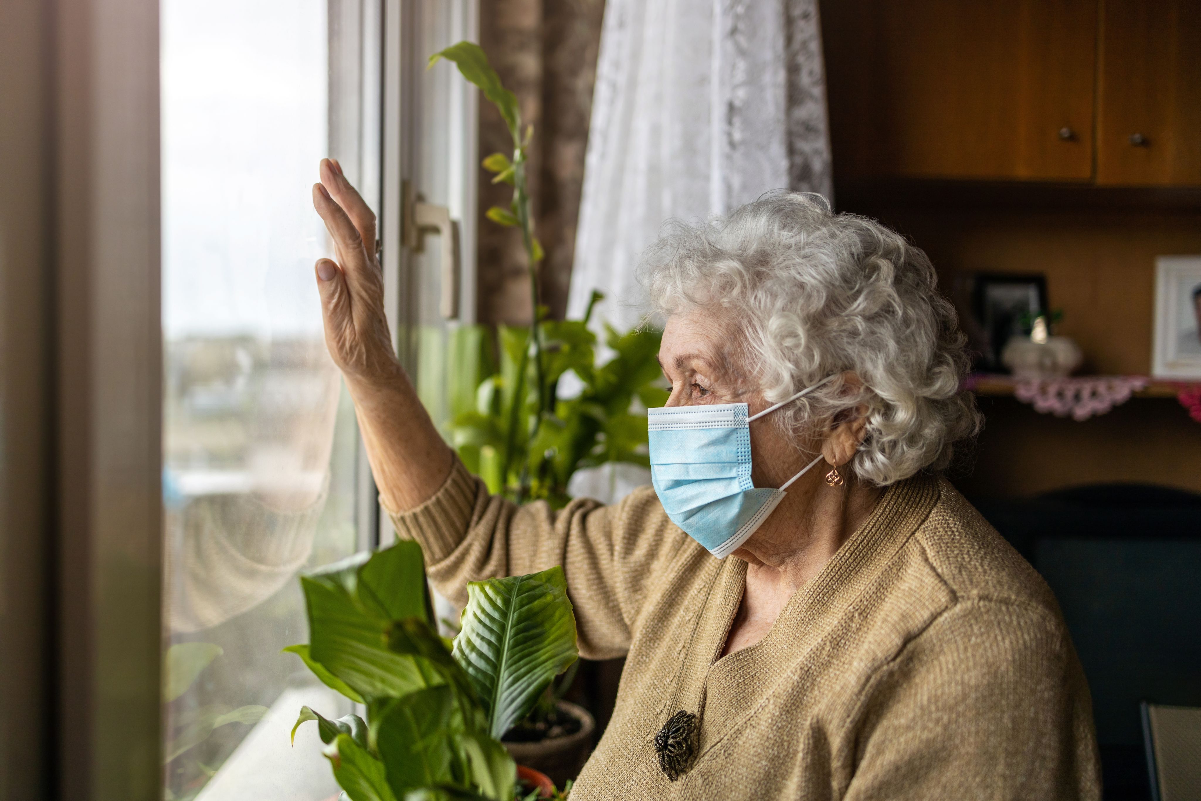 An elderly woman, wearing a clinical face mask, looks sadly out of a window