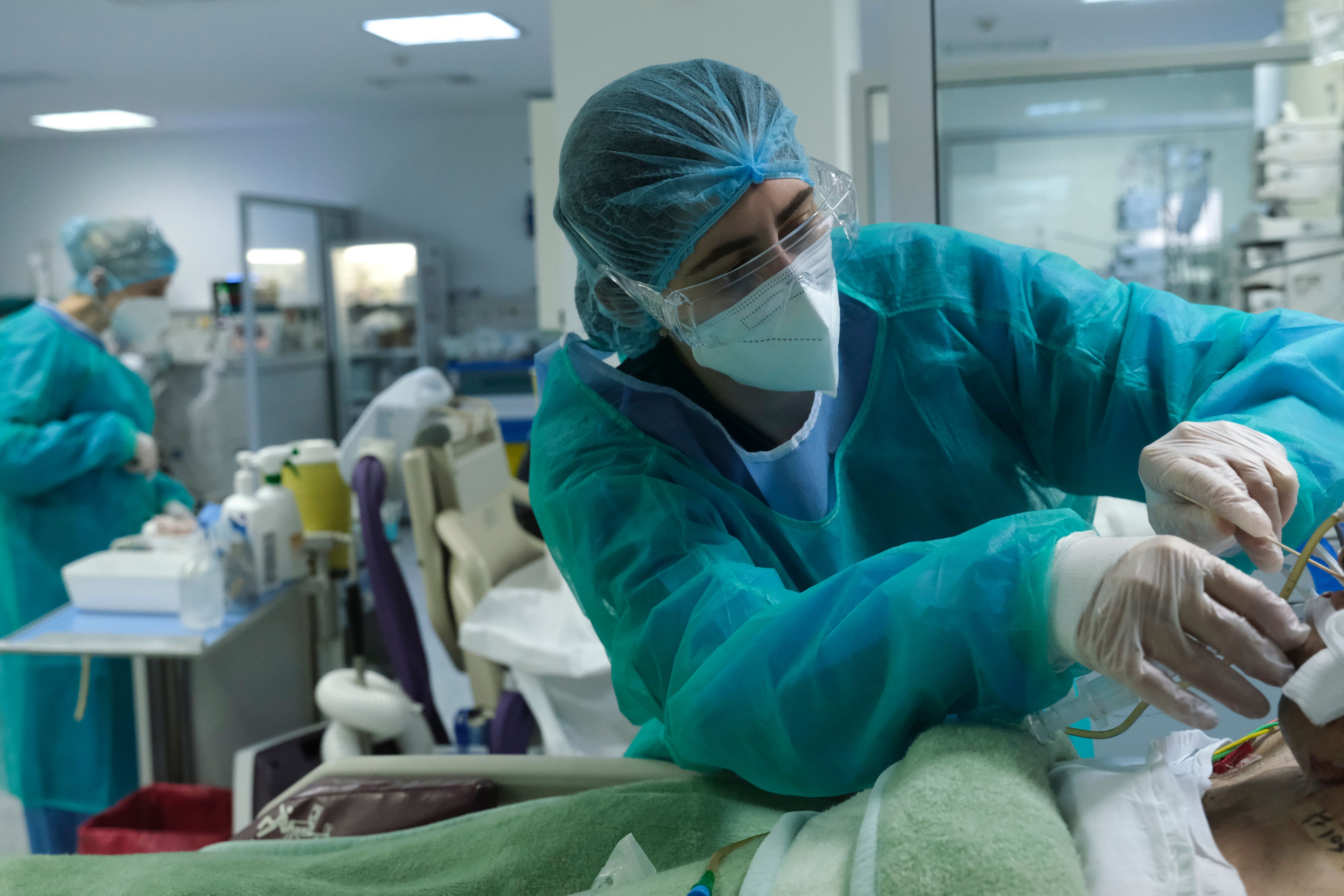 A medical professional wearing protective clothing and face mask attends to a patient in a busy hospital