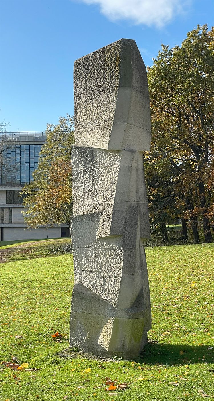The Standing Stone sculpture on a grassy bank. Made from rough stone, its four triangular prisms are stacked on top of one another, each twisted round from the last and partially sunken into the stone below.