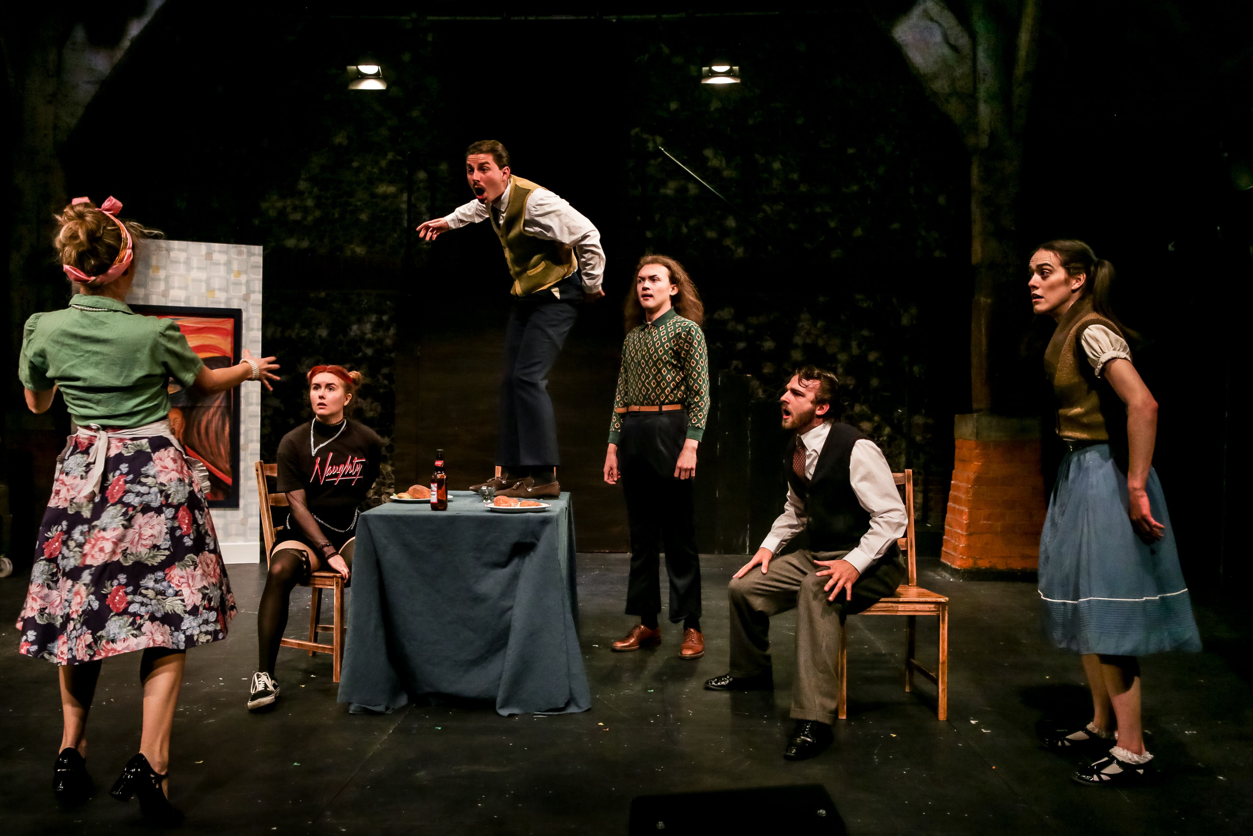Six actors, wearing a mixture of vintage 20th century costumes on stage, one standing on a table and gesticulating wildly.