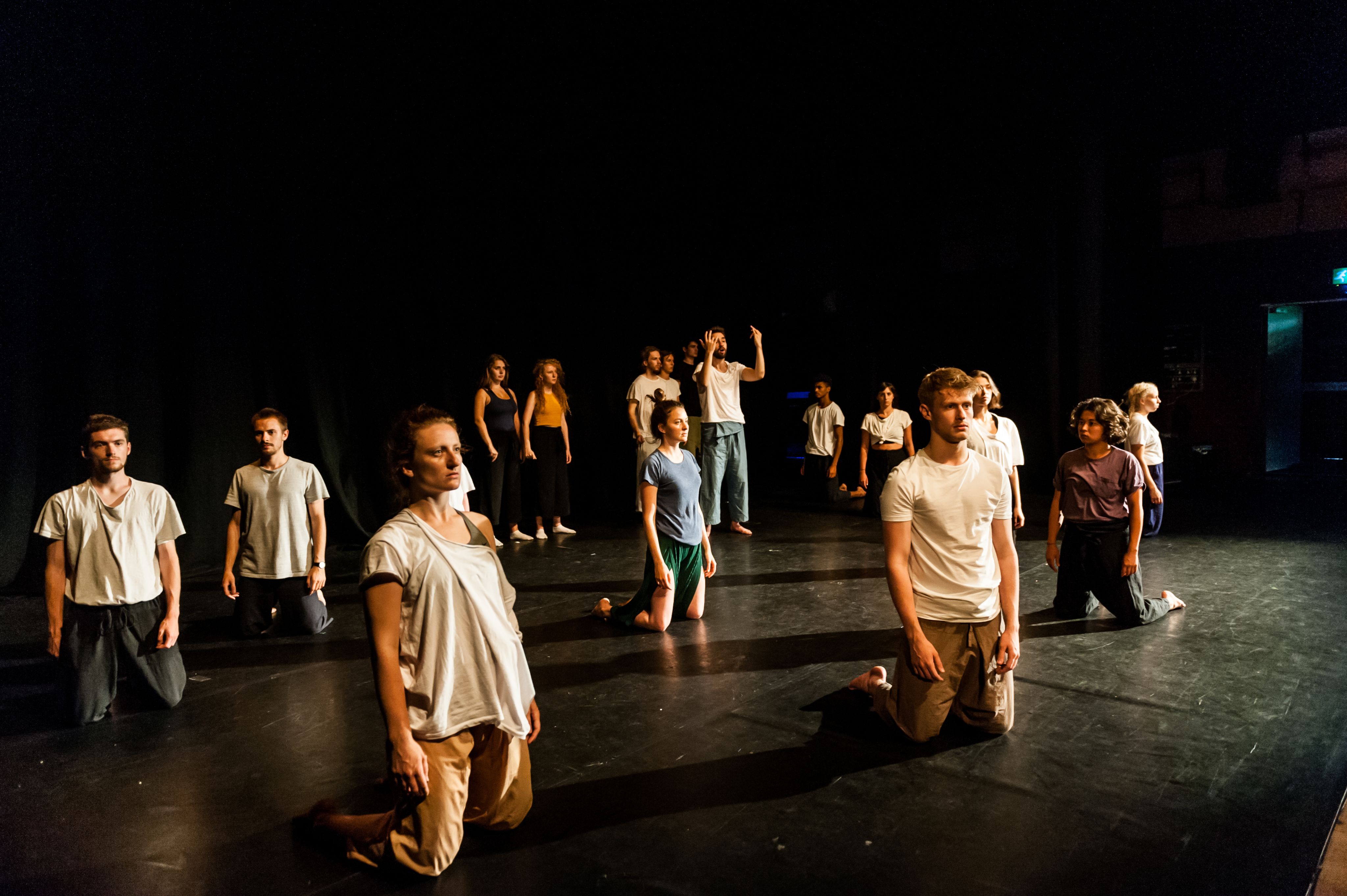 A group of young people, men and women, some kneeling and some standing, on a theatre stage, most looking dejected.