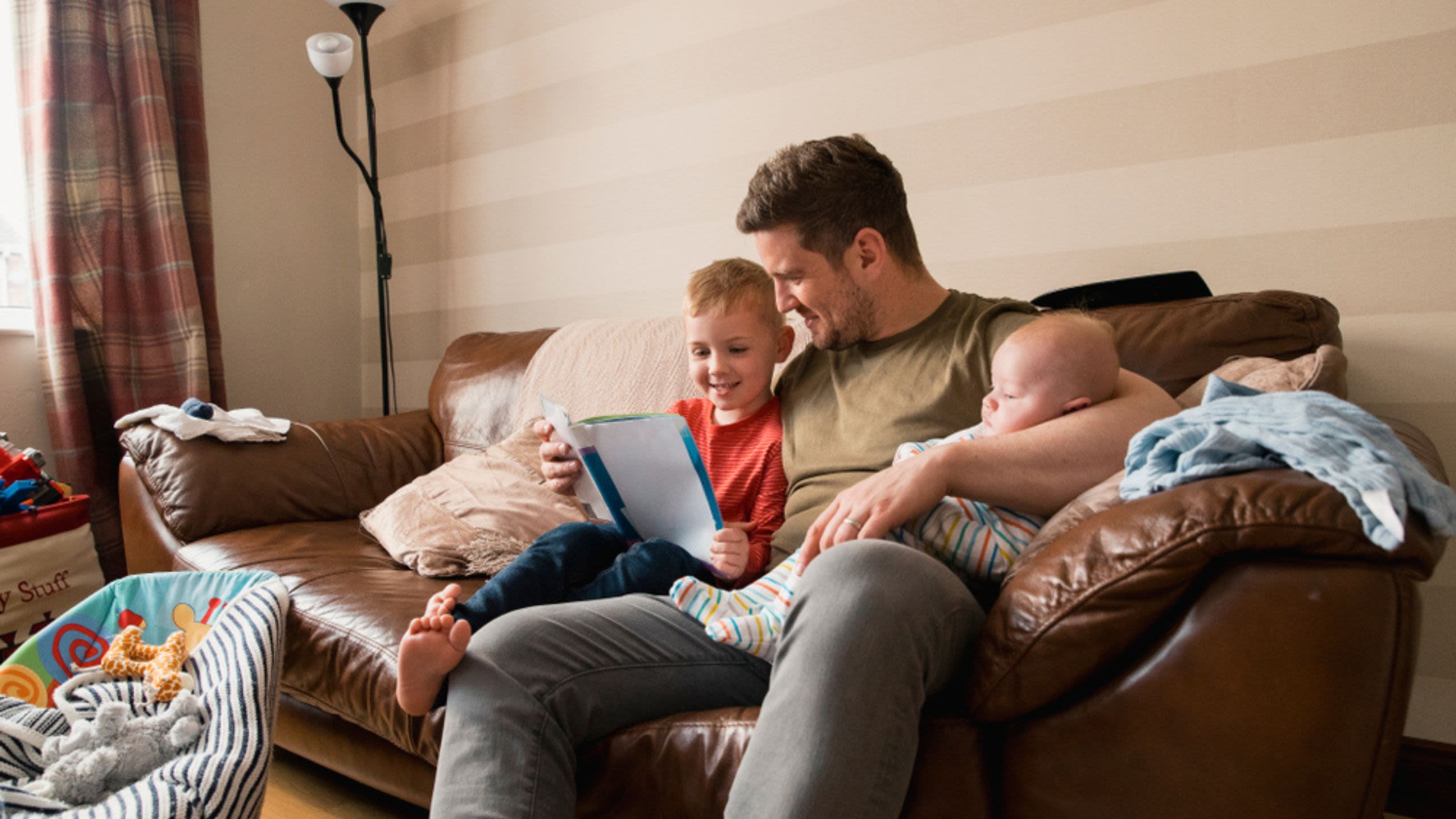 Father on sofa with baby and readying book to toddler