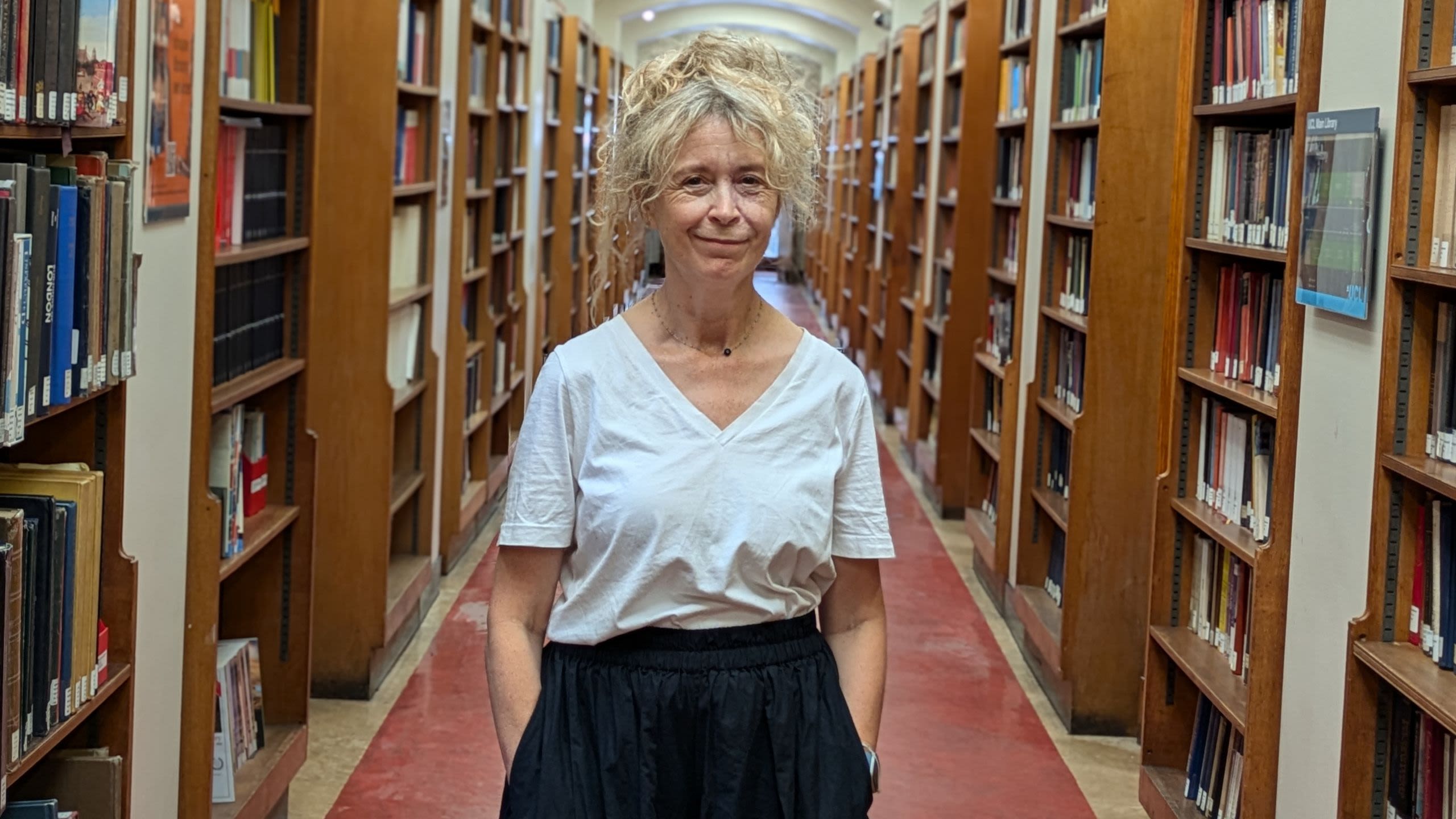 Portrait of Professor Lucy Noakes, standing in a corridor lined with bookshelves, with her hands in her pockets and looking relaxed