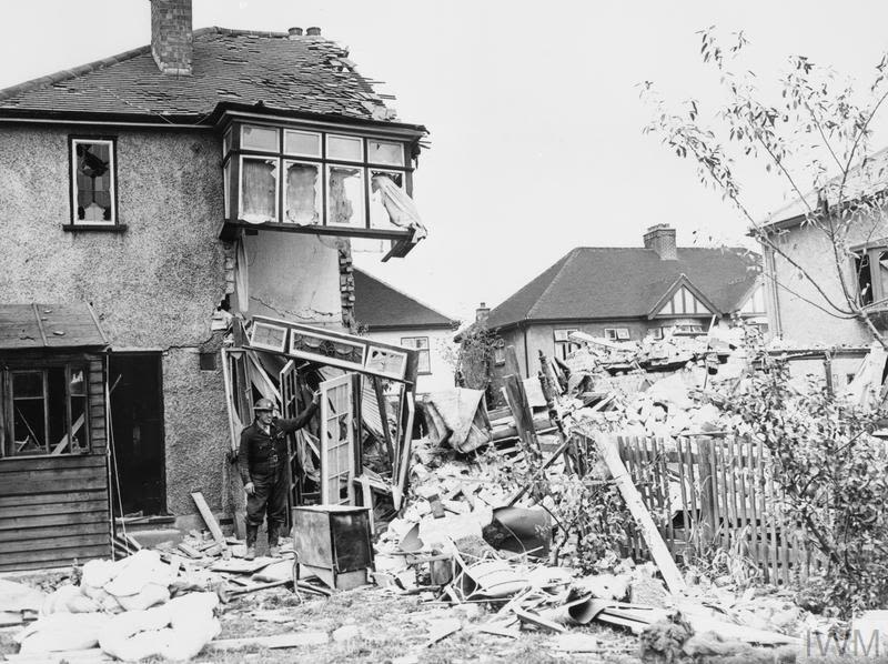 Archive photo of a man, wearing a tin hat and overalls, standing next to a semi-destroyed house and a pile of rubble, courtesy of the Imperial War Museum