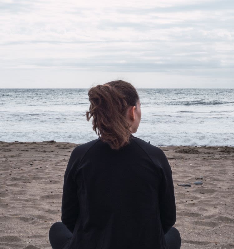 A young woman sits, with her back to the camera, on a beach looking out to sea