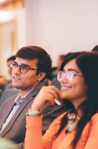 A man and a woman, both wearing glasses and looking ahead at an unseen speaker, sit together at a conference