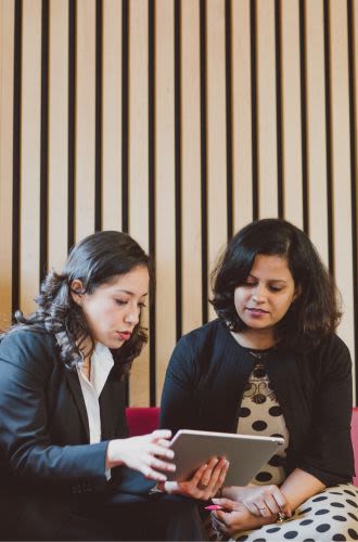 Two professionally-dressed women sit chatting whilst looking at something on a tablet