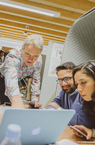 A male teacher leans forward and points at something on a laptop while while two students look on