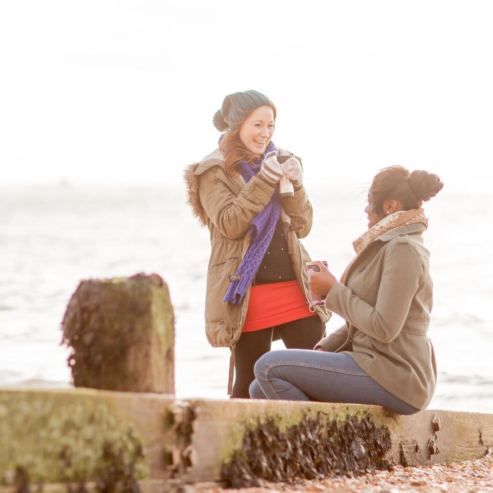 Two young women, wearing winter clothes, sit on a beach chatting on a cold day