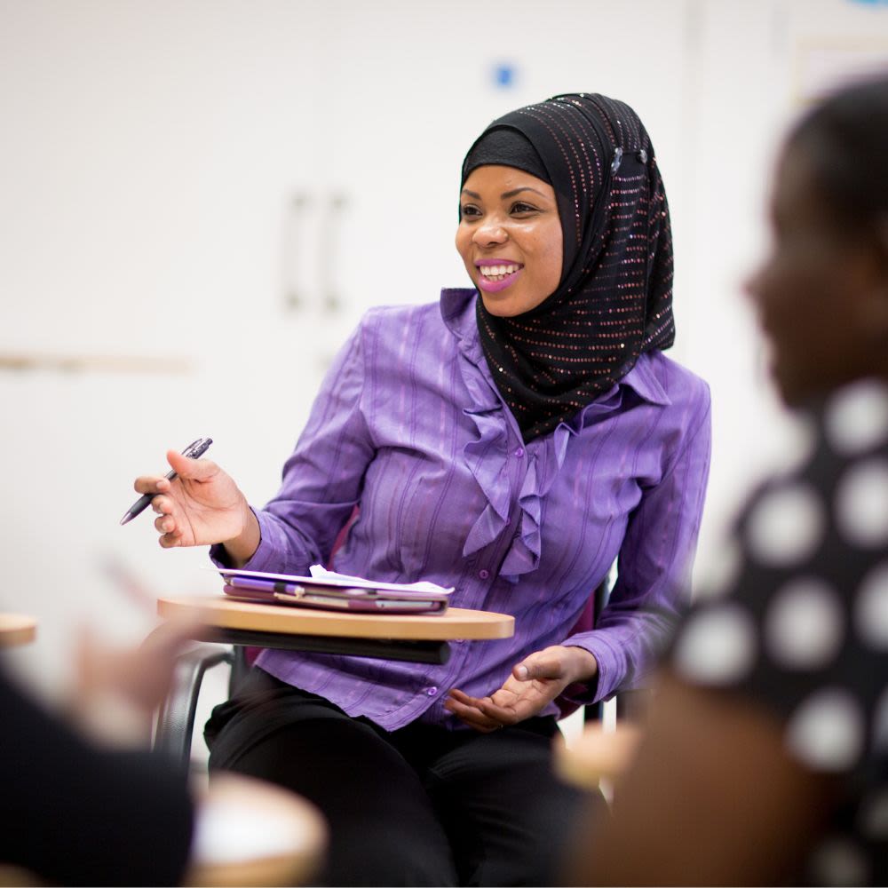 A young professional woman, wearing a headscarf, sits in a classroom environment in conversation with someone unseen