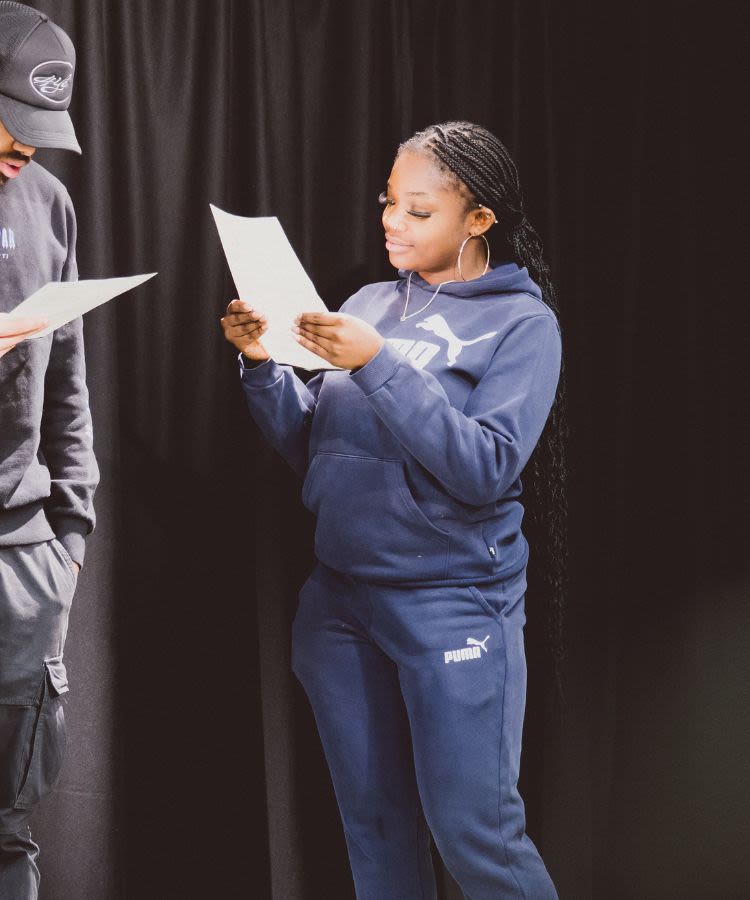 A young woman of colour stands in a theatre studio studying lines on a sheet of paper