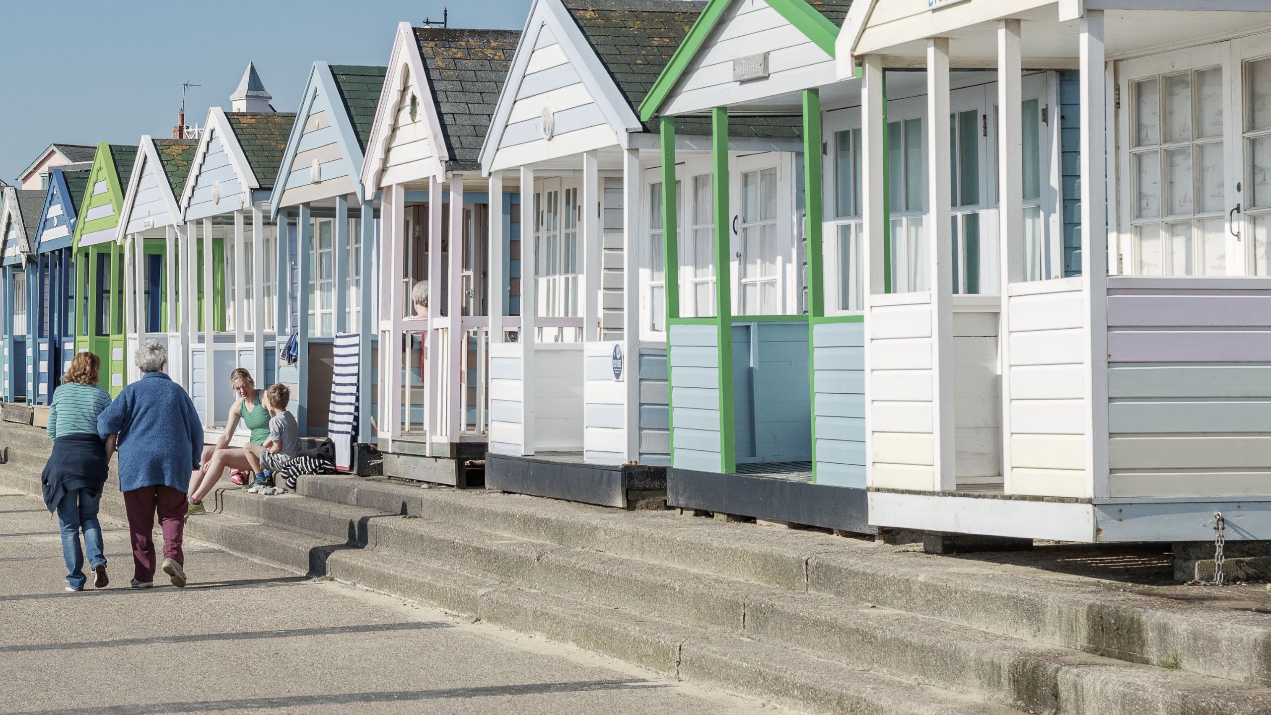 A couple of women walk along a seafront promenade beside a row of beach huts