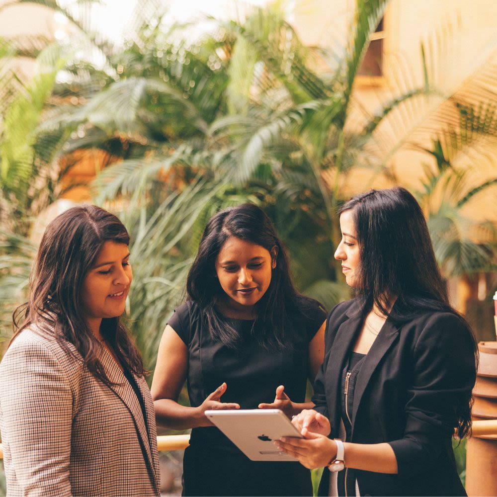 Three professional young women stand chatting, whilst one holds up a tablet