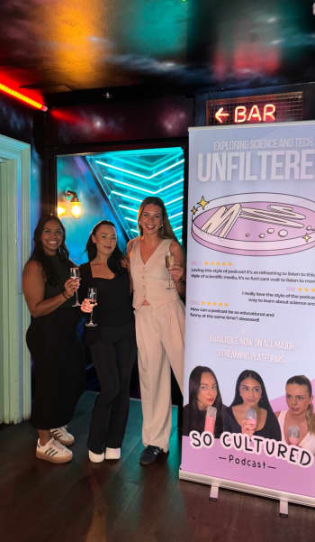 The three hosts of So Cultured, including Liv, stood next to a banner promoting the podcast